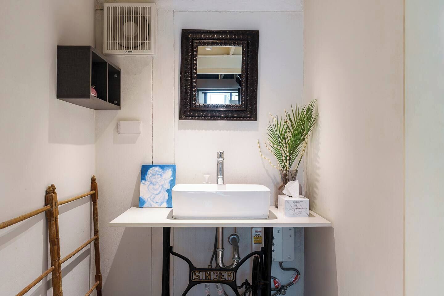Une salle de bain avec un lavabo blanc, un miroir, un radiateur, un distributeur de serviettes et une plante.