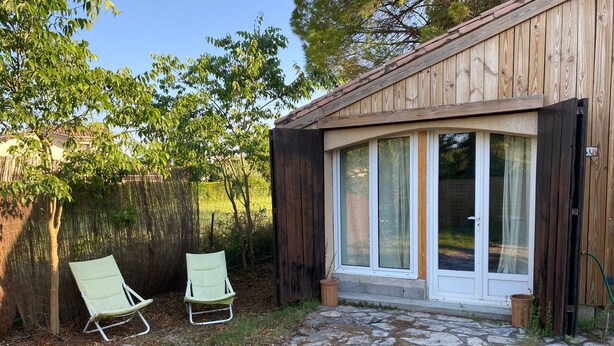 A wooden building with a stone patio, two green chairs, and a glass door. - Welcome to the Duchy of Uzes, in the charming village of Ste Eulalie. (Garrigues-Sainte-Eulalie)