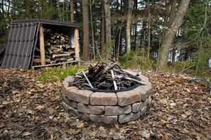 A stone fire pit surrounded by dry leaves, a wooden shed with logs, and a forested area. - Rosseau Cathedral Grove (Rosseau)