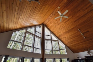 A room with a sloped wooden ceiling, a ceiling fan, large windows with a view of trees, and a wooden cabinet. - Rosseau Cathedral Grove (Rosseau)