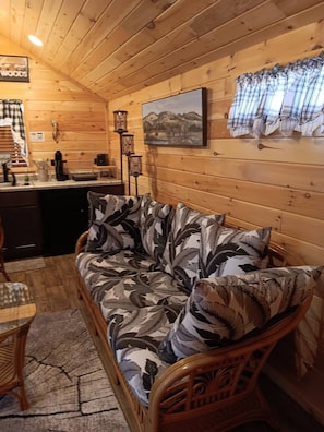 Un acogedor interior de cabaña con una pared de madera, un sofá con cojines estampados, una zona de comedor con mesa y sillas, y un cuadro sobre el sofá.