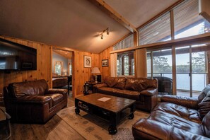 Ein Holzinterieur mit Ledermöbeln, einem Flachbildfernseher und großen Fenstern mit Blick auf ein Gewässer.