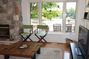Ein Wohnzimmer mit Steinwand, Kamin, Fernseher und Blick auf eine Terrasse und Bäume durch große Fenster.