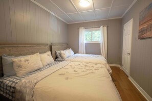 A bedroom with two beds, a window with curtains, a door, and a painting on the wall. - Steps To Lake Joe Ontario (Lake Joseph)