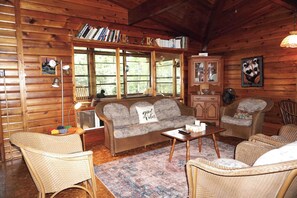 Ein gemütliches Blockhaus-Interieur mit einer Couch, Sesseln, einem Bücherregal und einem Fenster mit Blick auf Bäume.