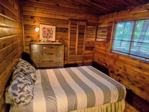 Ein Zimmer in einer Holzhütte mit einem Bett, einem Schminktisch, einem gerahmten Bild und einem Fenster.