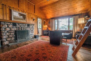A wooden cabin interior with a stone fireplace, a sofa, a ladder, and a large window.