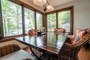 A dining area with a large table and wicker chairs, large windows with a view of trees, and a hanging light fixture.