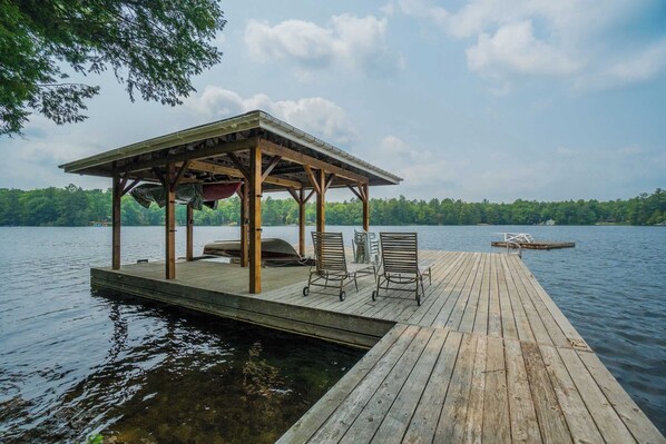 A wooden dock extending into a lake, with a covered seating area featuring chairs and a grill.