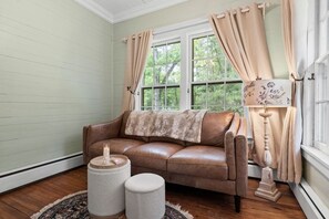 A room with a brown leather sofa, a round ottoman, a lamp with a floral shade, and a window with beige curtains. - The Rourk House | Historic 1927 Southern Home (Shallotte)