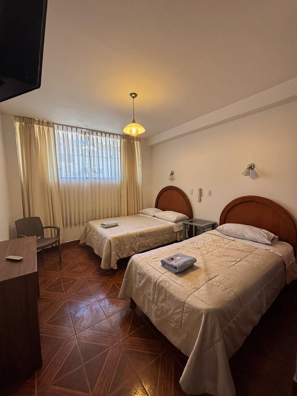Habitación de hotel con dos camas, un escritorio, una silla, un televisor, una ventana con cortinas y una lámpara colgante.