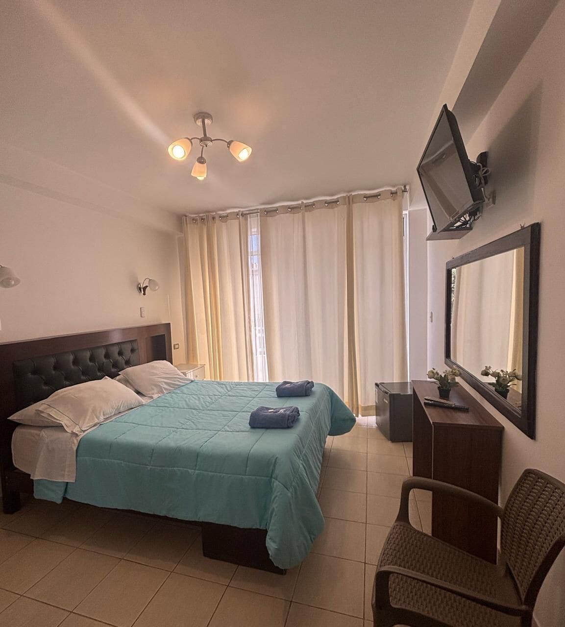 Habitación de hotel con cama, escritorio, silla, televisor y ventana con cortinas.