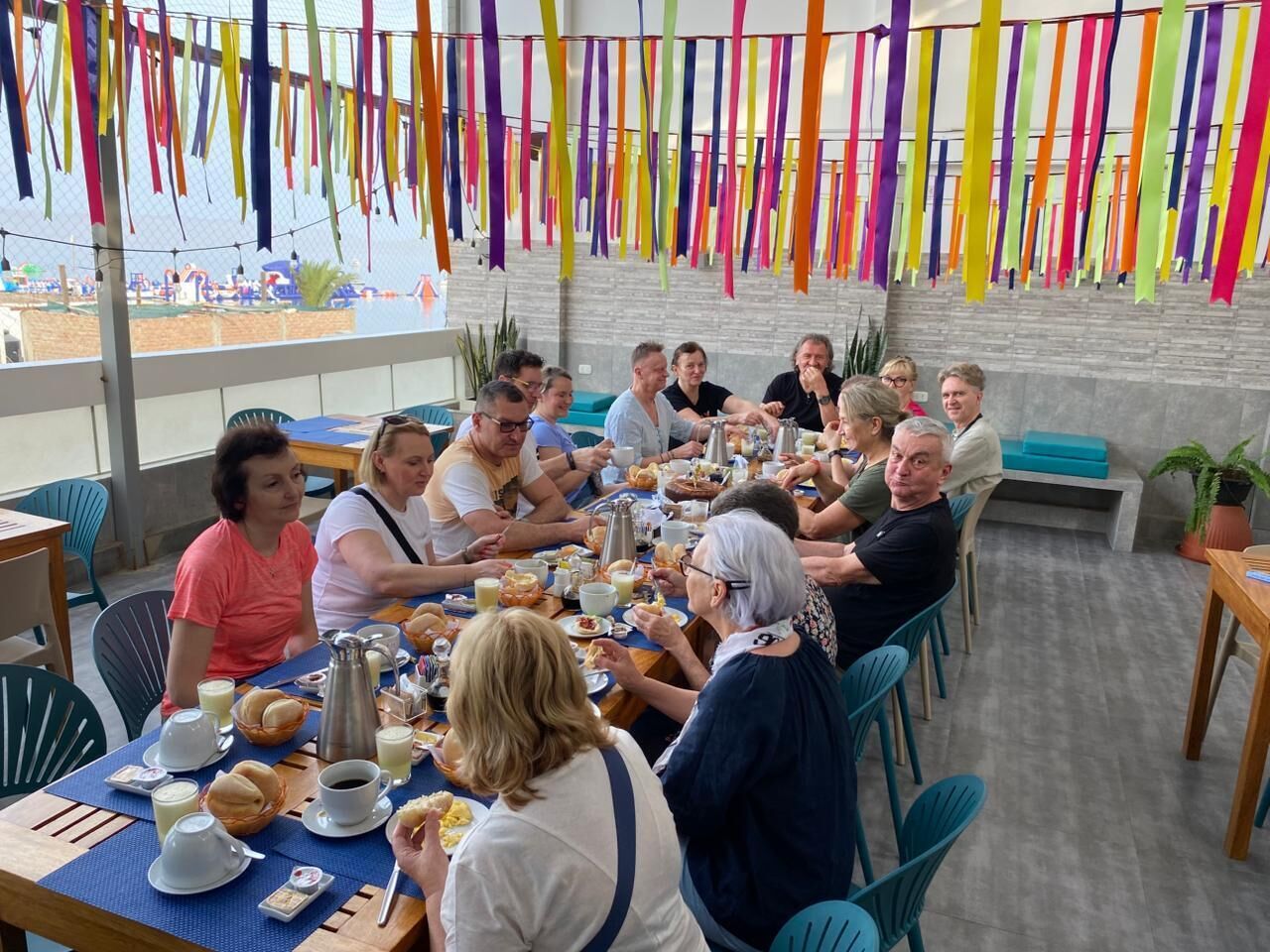 Un grupo de personas cenando en una mesa con vistas a la playa, rodeado de decoraciones colgantes de colores.