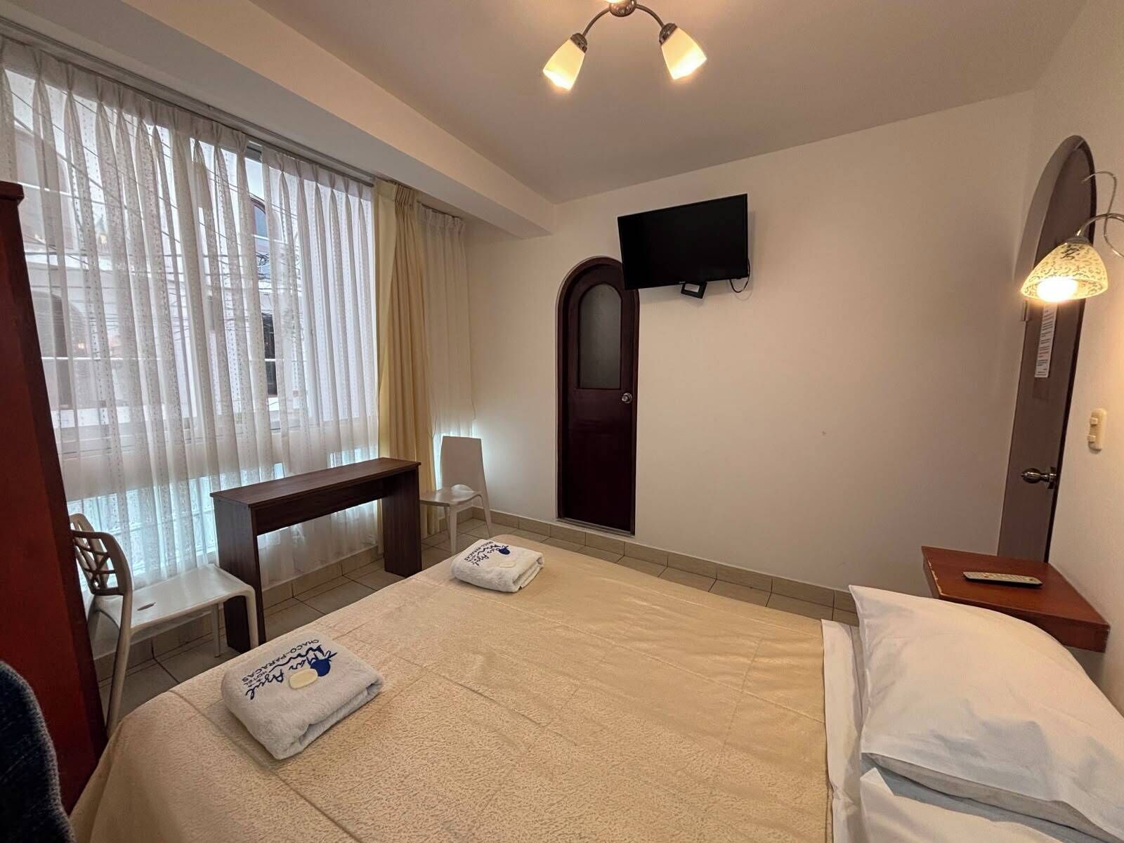 A hotel room with a bed, a desk, a chair, a TV, and a window with curtains.