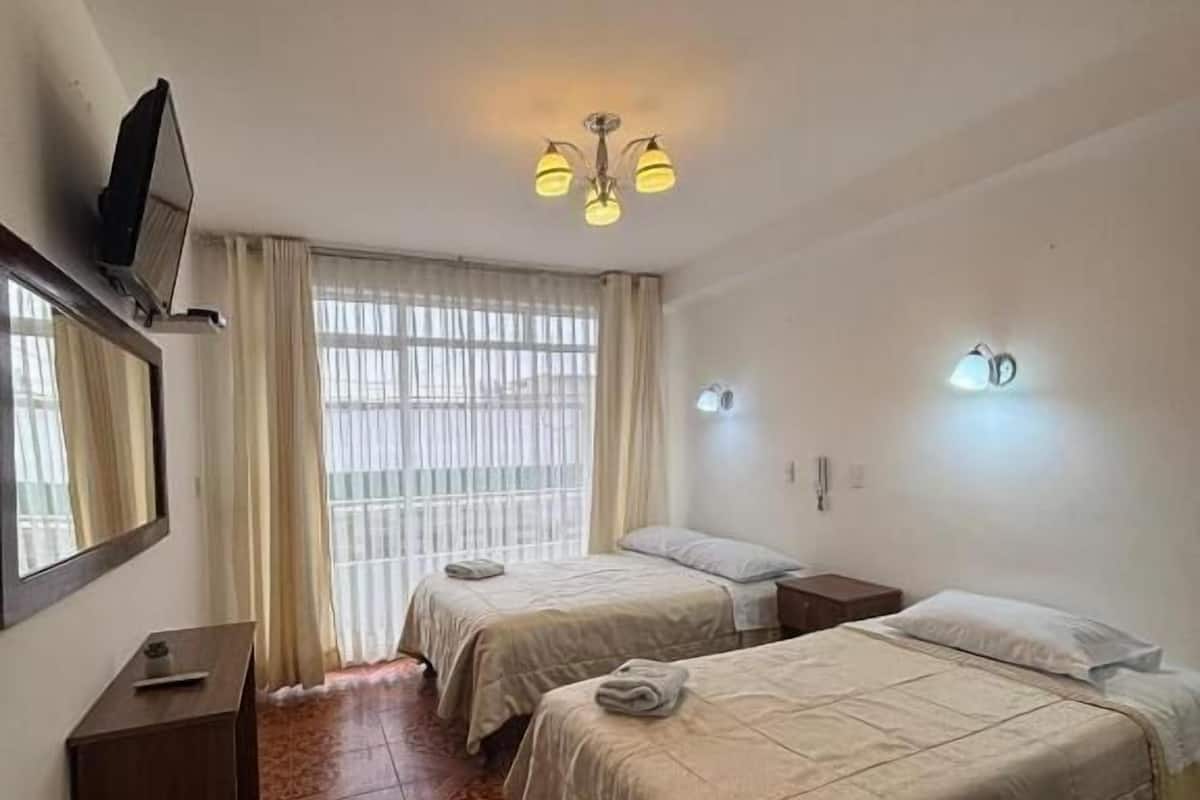 A hotel room with two beds, a television, a ceiling fan, a window with curtains, and patterned floor tiles.