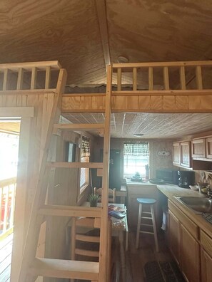 Un dormitorio tipo loft con una escalera de madera que conduce a una cama individual, una kitchenette con fregadero y estufa, y un ventanal con cortinas.