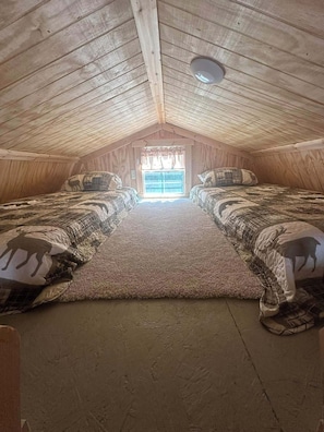 A cozy attic bedroom with a sloped ceiling, two beds with patterned bedding, a large window, and a ceiling light.