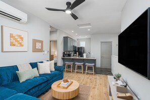 Ein modernes Wohnzimmer mit einem blauen Sofa, einem runden Couchtisch, einem Deckenventilator und einer Küchenzeile mit Barhocker.