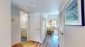 Un couloir avec une porte donnant sur une salle de bain, une chaise en osier et un tableau encadré au mur.