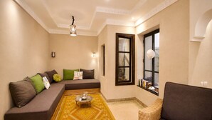 A living room with a sofa, a small table, a wicker chair, a patterned rug, and a window with books on the sill. - 2-bedroom riad in enchanting Marrakech with cool AC breeze (Marrakech)
