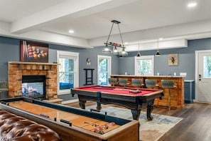 Uma mesa de bilhar com feltro vermelho, uma mesa de futebol de mesa e um bar com banquetas.