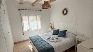 A bedroom with a bed, a bedside table, a dresser, a window with curtains, a hanging light fixture, and a wall decoration.
