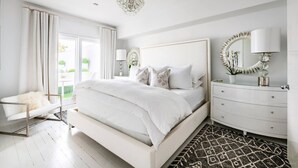 Ein helles Schlafzimmer mit einem großen Bett, einem Schminktisch, einem Sessel und einer Deckenleuchte.