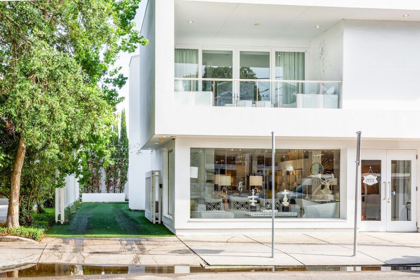 A modern two-story building with a glass facade, a visible interior with furniture, and a green lawn in front.