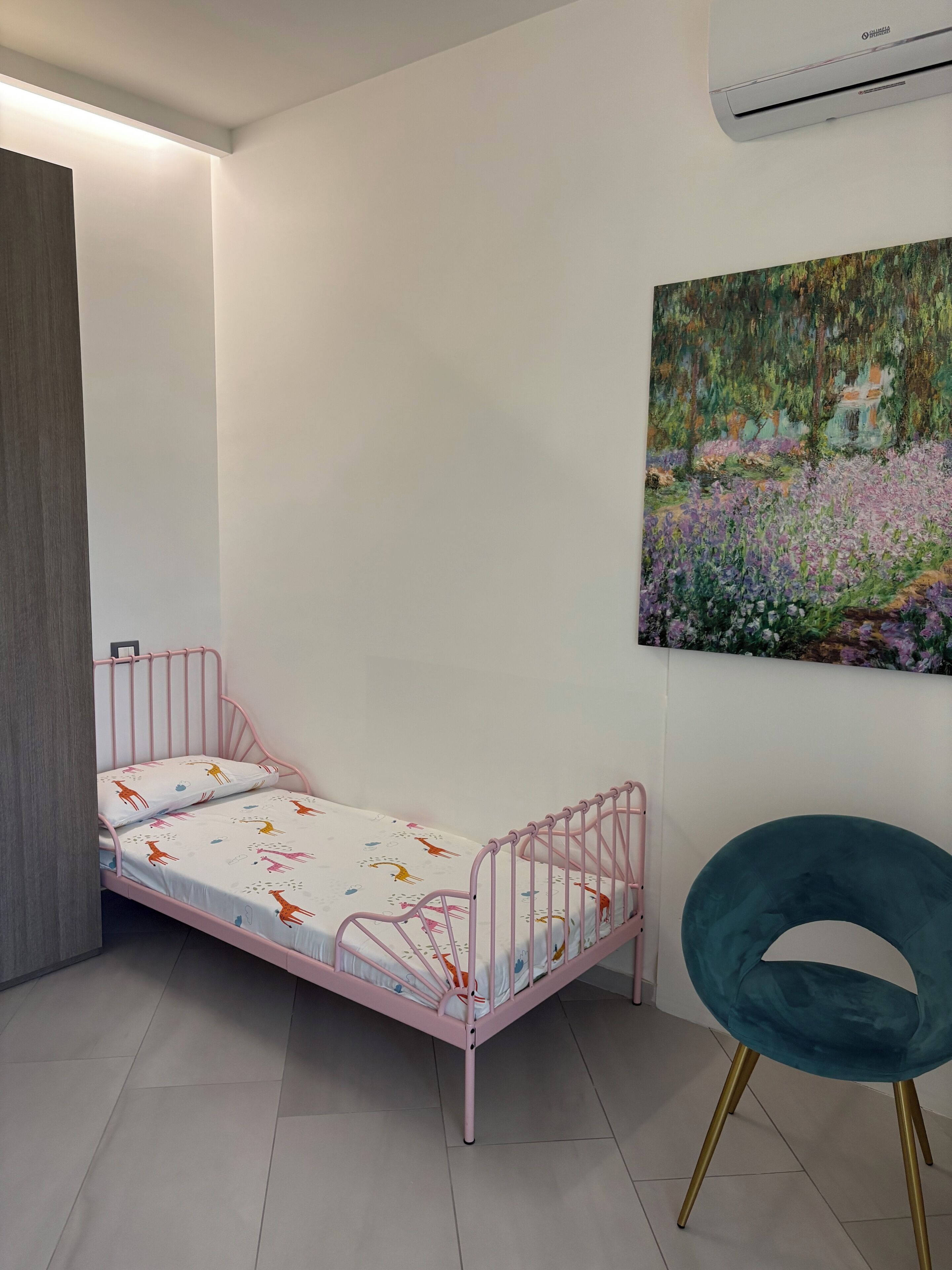 Ein kleines Zimmer mit einem pinkfarbenen Metallbett, einem türkisfarbenen Sessel und einem Gartenbild an der Wand.