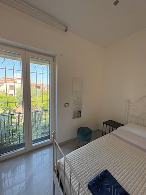 Ein Schlafzimmer mit einem Bett, einem kleinen Hocker, einem Nachttisch und Blick durch eine Schiebetür.