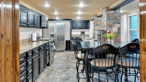 Una cocina con armarios oscuros, un refrigerador de acero inoxidable, una encimera de granito y un comedor con mesa y sillas.