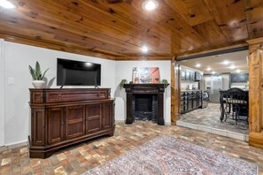 Ein Wohnzimmer mit Kamin, Fernseher und einer durchgehenden Tür erkennbaren Küche.