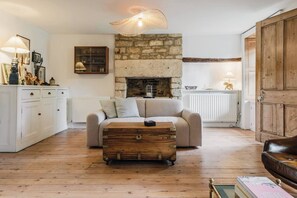 A cozy living room with a stone fireplace, a sofa, a wooden chest, and a white cabinet. - Cotswolds Cottage - Garden & Ground Floor Bedroom (Milton-under-Wychwood)