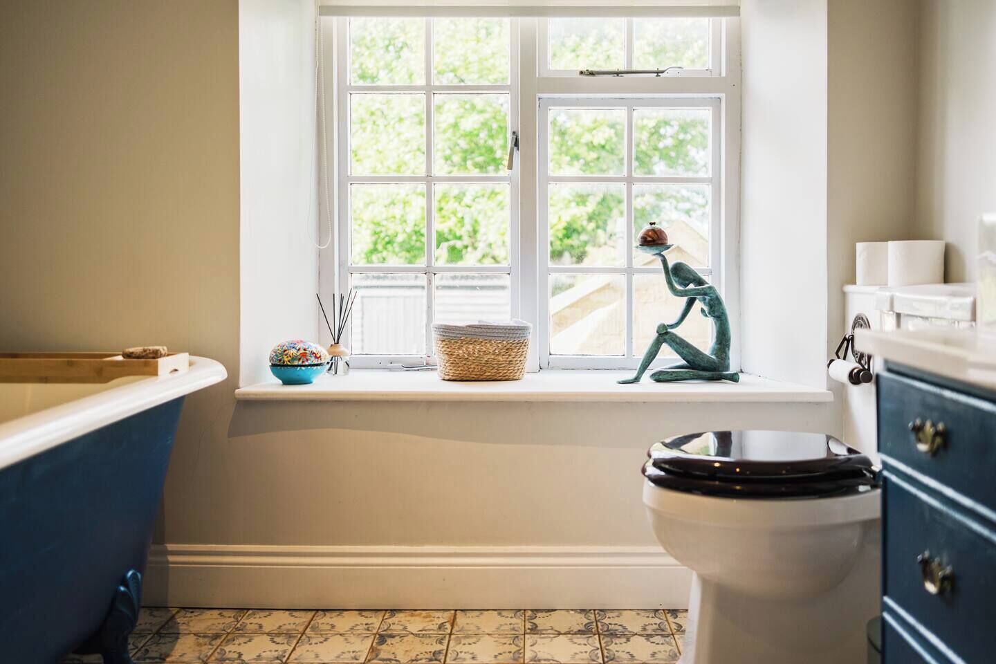 A bathroom with a bathtub, a toilet, a window with a view of greenery, and a decorative sculpture on the windowsill.