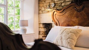 Una camera da letto con parete in pietra, una testiera in legno, un letto bianco con cuscino decorato, una lampada su un comodino e una finestra affacciata sul verde.