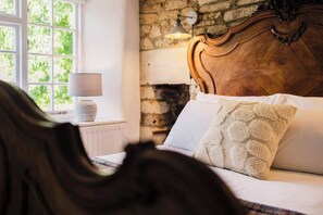 A bedroom with a stone wall, a wooden headboard, a white bed with a textured pillow, a lamp on a bedside table, and a window with greenery outside. - Cotswolds Cottage - Garden & Ground Floor Bedroom (Milton-under-Wychwood)