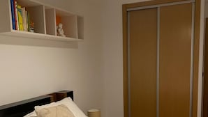 En snyggt bäddad säng med vita sängkläder och en beige kudde, ett sängbord med en lampa, en hylla med böcker och en garderob med skjutdörrar.