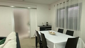 En matplats med ett bord för fyra personer, en soffa, ett litet skåp med dekorativa föremål och ett fönster med persienner.