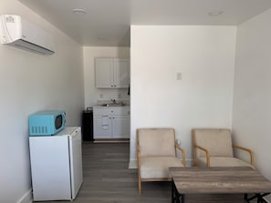 A compact kitchen with white cabinets, a sink, and a microwave on a white appliance. There are two beige chairs and a wooden table in the living area.