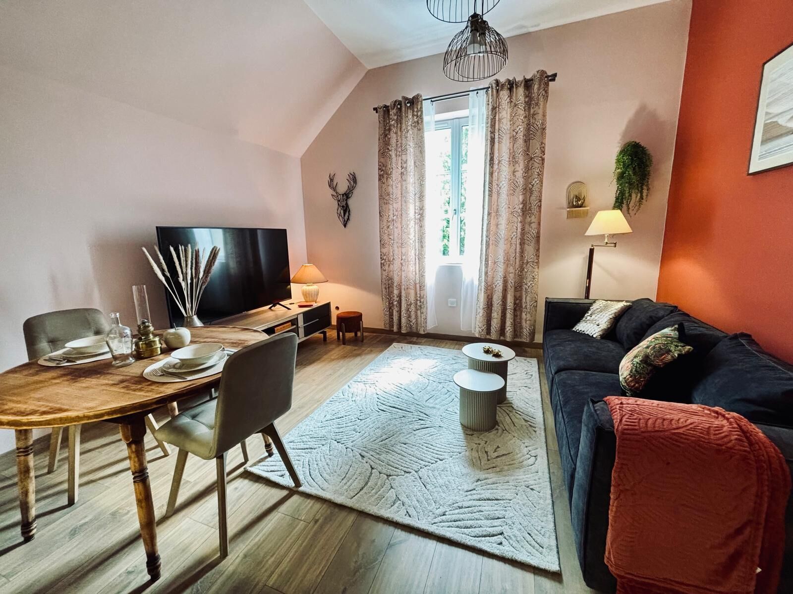 Ein Wohnzimmer mit Esstisch, Sofa, Fernseher und einem großen Fenster mit Vorhängen.