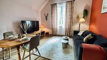 Ein Wohnzimmer mit Esstisch, Sofa, Fernseher und einem großen Fenster mit Vorhängen.