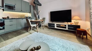 En moderne stue med et fladskærms-tv, et spiseområde med et bord og stole, og et køkken med grå skabe og en mikrobølgeovn.