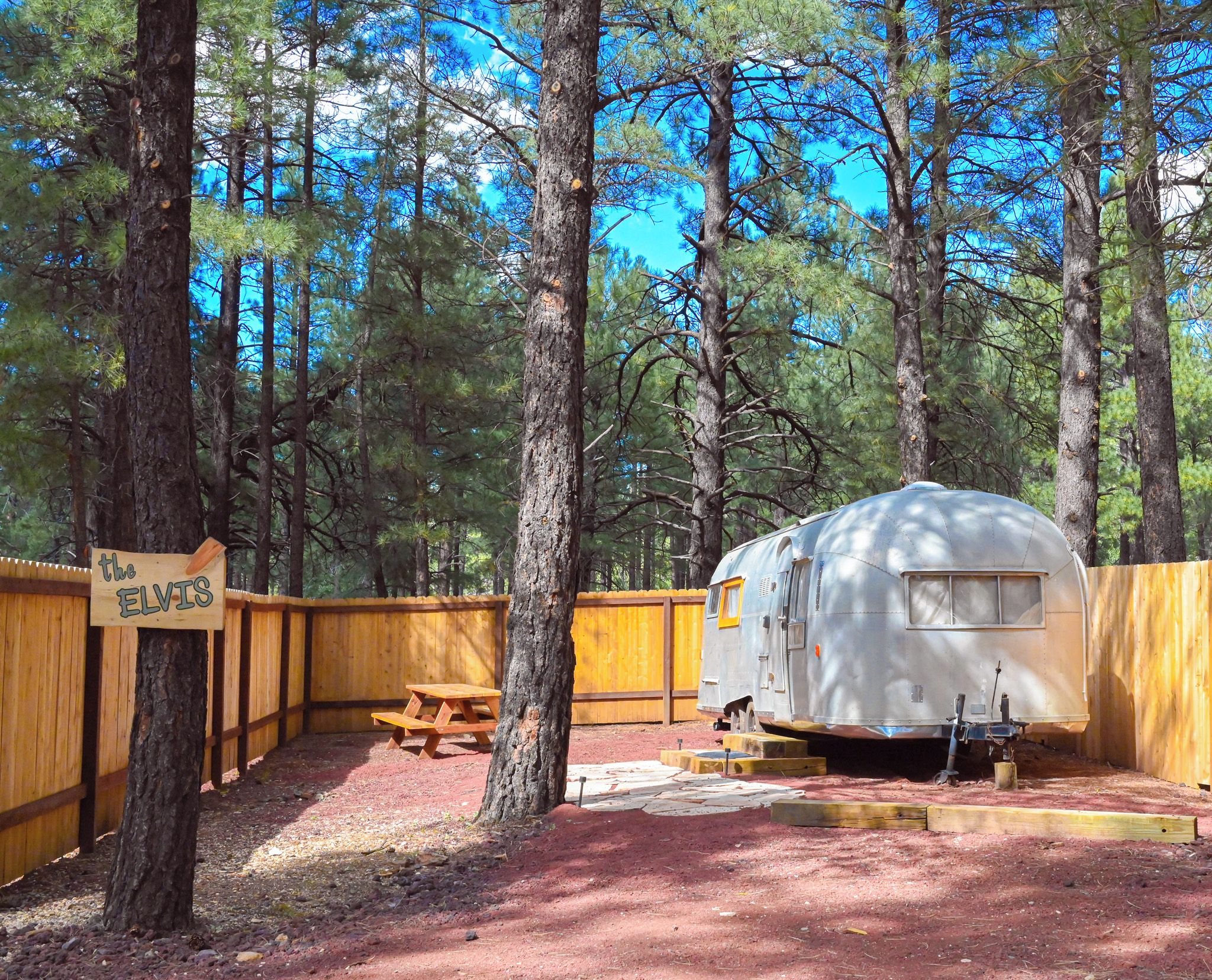 Un campamento con cerca de madera, una mesa de picnic y una caravana clásica estacionada en un área boscosa.