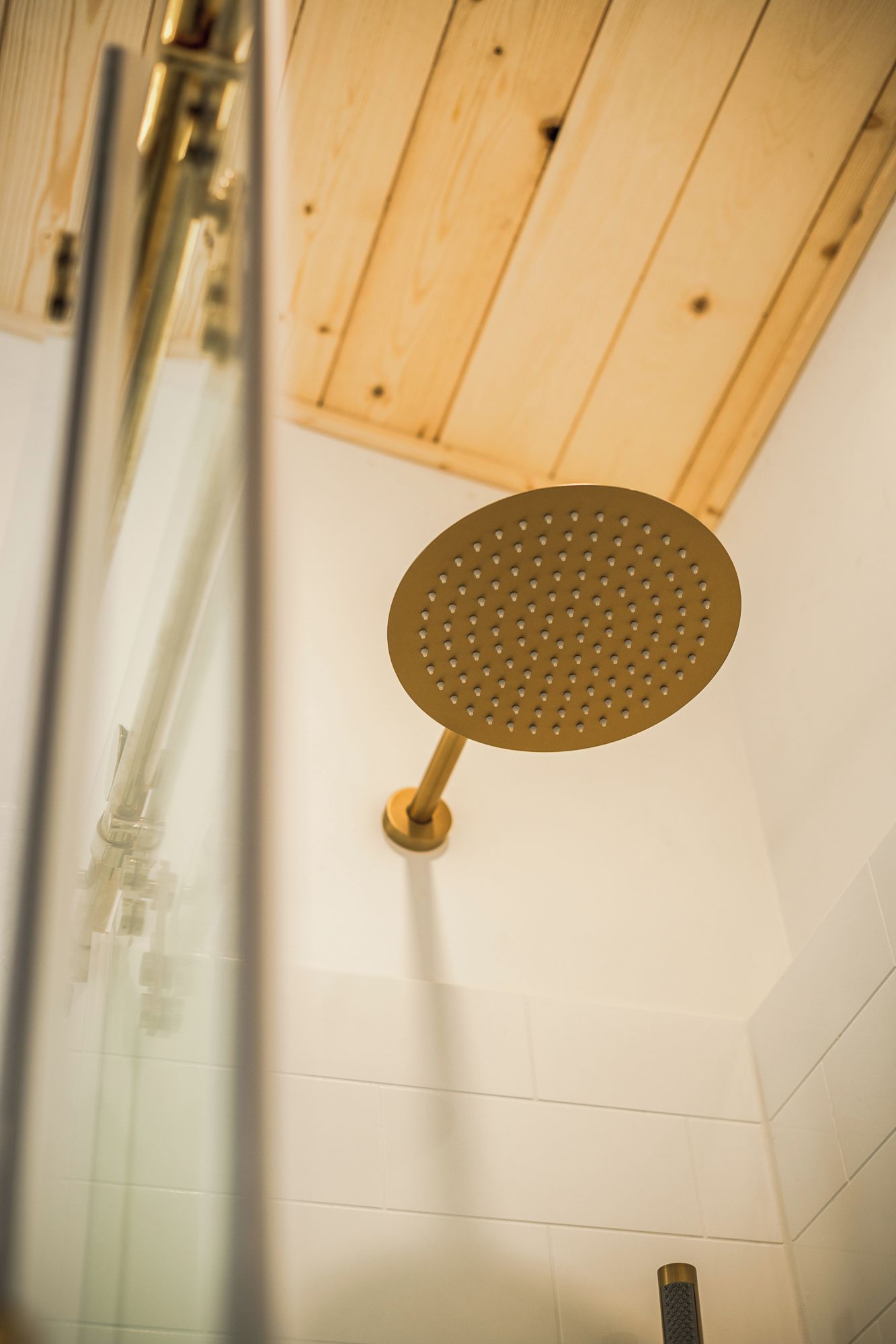 Un soffione dal design circolare forato, fissato a una parete con soffitto in legno e piastrelle bianche.