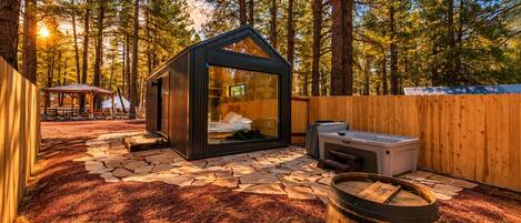 A cabin with a glass enclosure, a hot tub, and a stone-paved patio surrounded by trees.