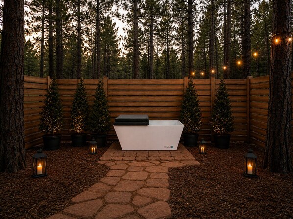 A wooden deck with a white bench, surrounded by pine trees and string lights.