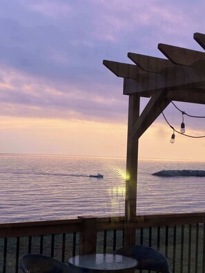 Eine Holzterrasse mit Tisch und Stühlen, mit Blick auf ein ruhiges Meer, ein Boot und einen Wellenbrecher.