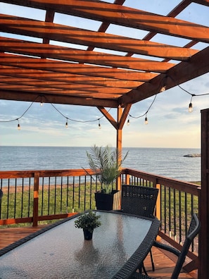 Ein ĂŒberdachter AuĂenbereich mit Tisch und StĂŒhlen, einem Blumentopf und Blick aufs Meer.