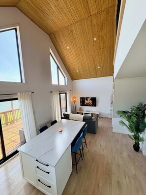 Amplia sala de estar con techo alto, una mesa de comedor, asientos, televisor y vistas al exterior.