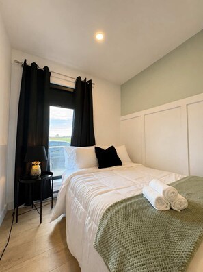 Un dormitorio con cama, una mesita de noche con lámpara, una ventana con cortinas y vistas al exterior.